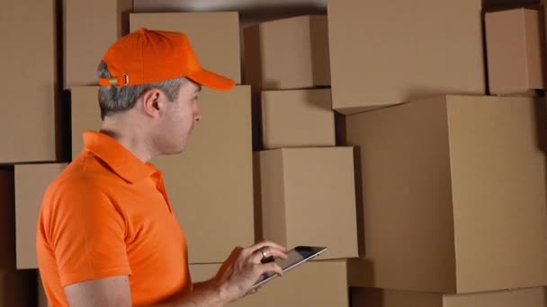 Trabajador de almacenamiento en uniforme naranja contando cajas y haciendo registros en su PC tableta contra cartones marrones. 4K video de estudio — Vídeo de stock