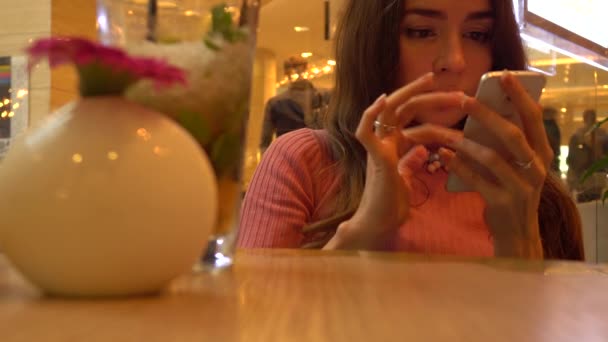 Mooie jonge vrouw in roze via haar telefoon in een café. Mobiele telefoon verslaving. 4k video — Stockvideo