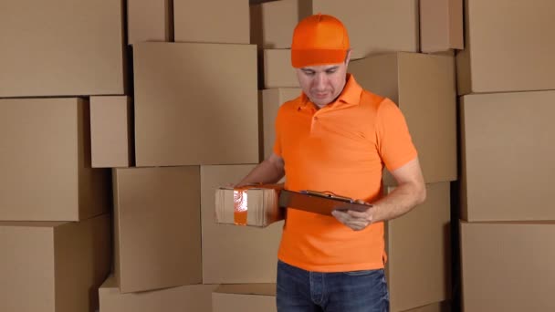 Hombre en uniforme naranja entregando paquete gravemente dañado al cliente. Fondo cartones marrones. Trabajo poco profesional y concepto de arrepentimiento. Disparo 4K — Vídeos de Stock