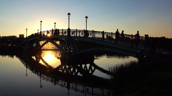 Siluetas de puente de parque arqueado y personas caminando al atardecer de verano — Foto de Stock