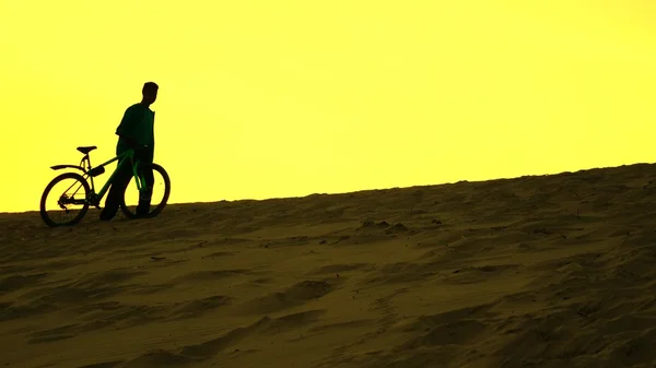 孤独骑自行车的人和他沿着沙丘，黄色日落颜色的自行车走的剪影 — 图库照片
