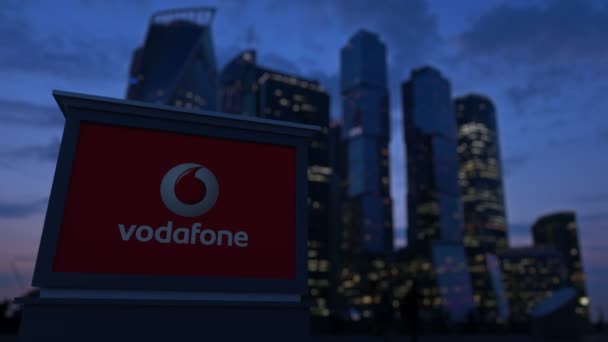 Gateskilt med Vodafone-logo om kvelden. Tåkete forretningsdistrikts skyskrapere bakgrunn. Redaksjonell 4K-klemme – stockvideo