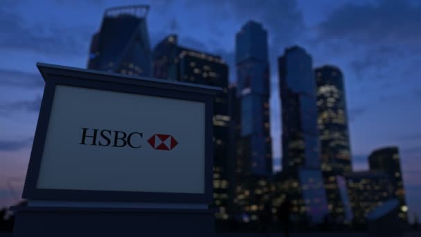 Уличная вывеска с логотипом HSBC вечером. Размытые данные о небоскребах в деловом районе. Клип в 4K — стоковое видео