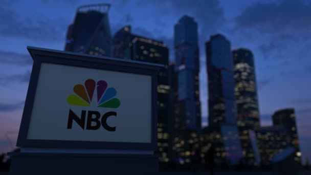 Уличная вывеска с логотипом Национальной вещательной компании NBC вечером. Размытые данные о небоскребах в деловом районе. Клип в 4K — стоковое видео