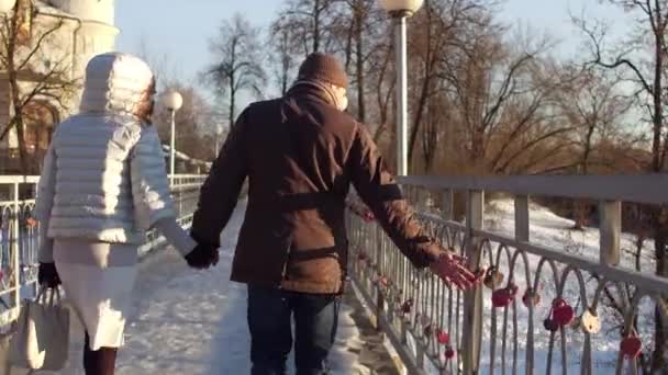 Мужчина и женщина ходят, держась за руки на маленьком мосту, полном замков любви. 4K steady icam shot, back view — стоковое видео