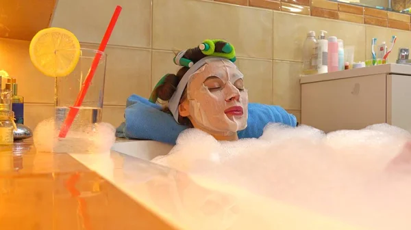 Красивая брюнетка в косметической маске расслабляется в пенной ванне. Косметические процедуры на дому — стоковое фото