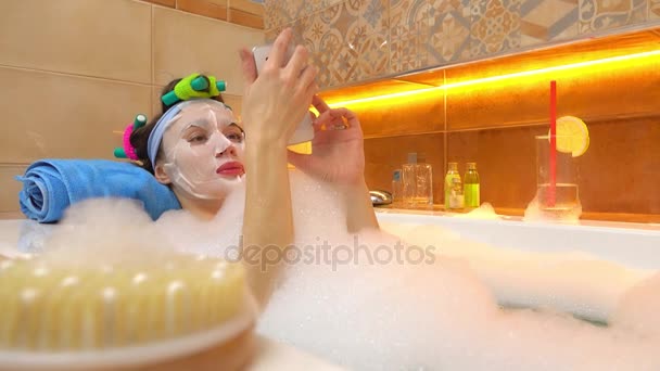 Brunette woman wearing face mask taps on her smartphone in foamy bathtub. 4K video — Stock Video