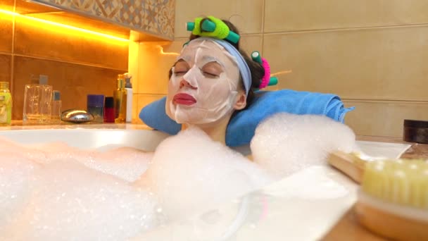 Bella giovane donna che indossa maschera cosmetica si rilassa nella vasca da bagno schiumosa. Trattamenti di bellezza a casa. Video 4K — Video Stock