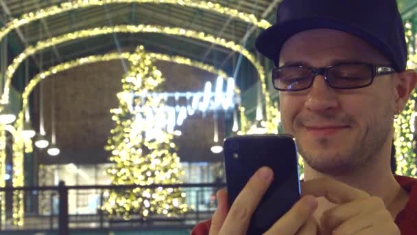 Ein glücklicher Mann mit schwarzer Randbrille blättert in einem weihnachtlich dekorierten Einkaufszentrum in seinem Handy. 4k-Video — Stockvideo
