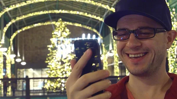 Happy programmer in black rim glasses answering video call on his mobile phone in a Cristmas decorated cafe — Stock Photo, Image