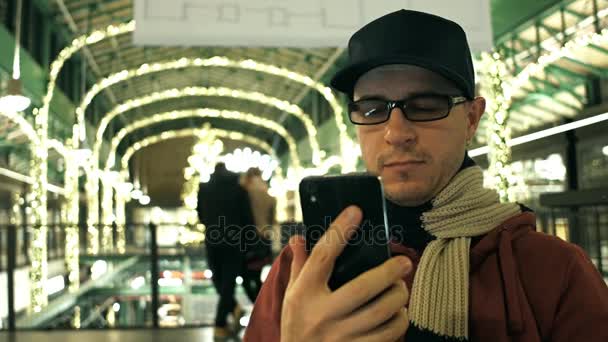 Uomo nerd in occhiali di bordo nero scorre app nel suo telefono cellulare in un centro commerciale. Video 4K — Video Stock