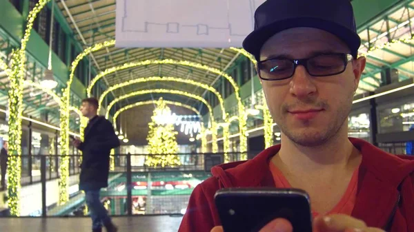 Homme aux lunettes de jante noires tapant un message sur son téléphone portable dans un centre commercial décoré de Noël — Photo