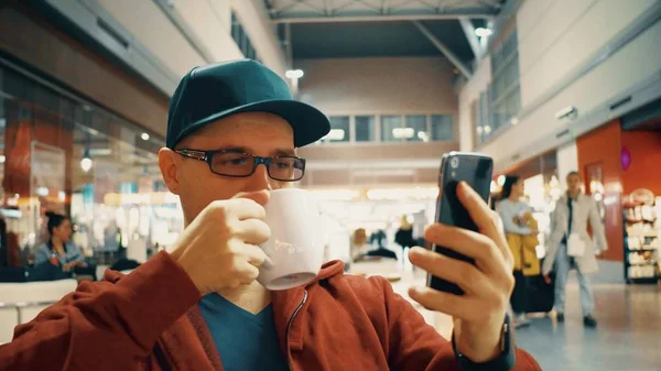 Homme aux lunettes de jante noires regardant la vidéo sur son téléphone portable et prenant une grande tasse de café au café de l'aéroport — Photo