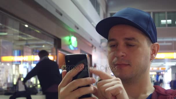 帽子を被った若い男は、空港のカフェで彼の携帯電話にアプリをスクロールします。4 k ビデオ — ストック動画