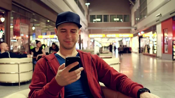Joven con gorra se desplaza aplicación en su teléfono inteligente en la cafetería del aeropuerto —  Fotos de Stock
