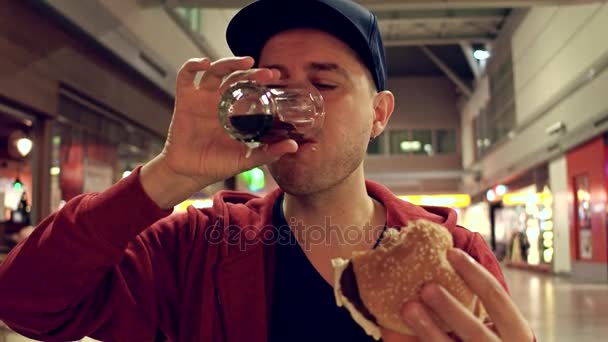 Junger Mann mit Schirmmütze isst Burger und trinkt Limonade im Flughafencafé. 4k-Video — Stockvideo