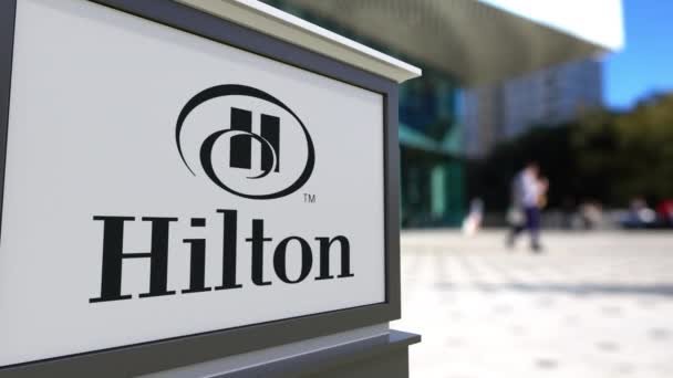 Placa de sinalização de rua com logotipo Hilton Hotels Resorts. Centro de escritórios desfocado e pessoas ambulantes fundo. Editorial 3D renderização 4K — Vídeo de Stock