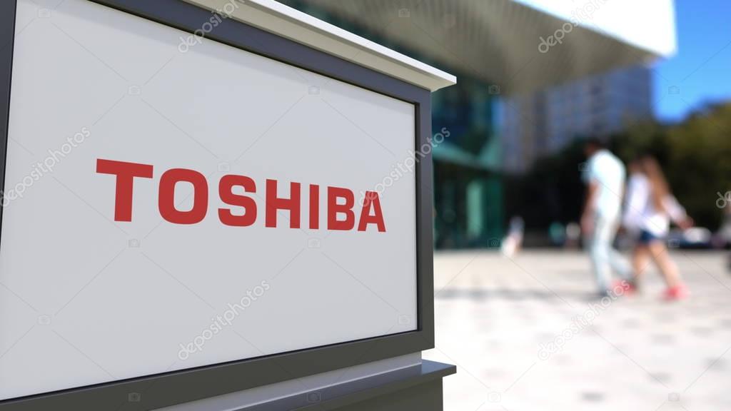 Street signage board with Toshiba Corporation logo. Blurred office center and walking people background. Editorial 3D United States