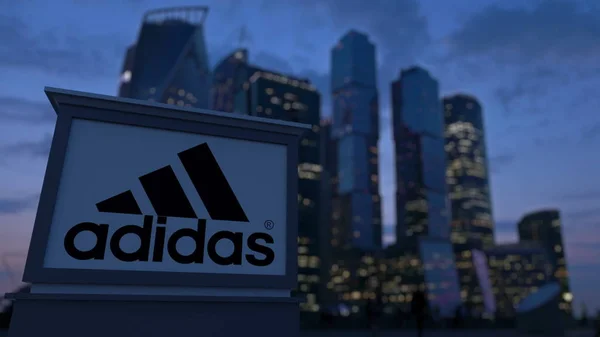 Papan nama jalan dengan tulisan dan logo Adidas di malam hari. Blurred bisnis daerah pencakar langit latar belakang. Perenderan 3D Editorial — Stok Foto