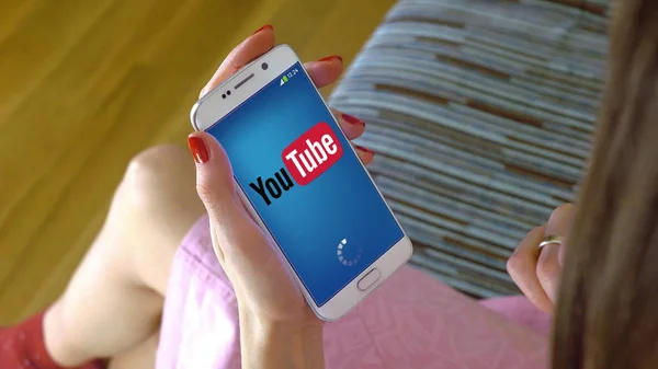 Молодая женщина держит мобильный телефон с загрузкой мобильного приложения YouTube. Концептуальная редакционная CGI — стоковое фото