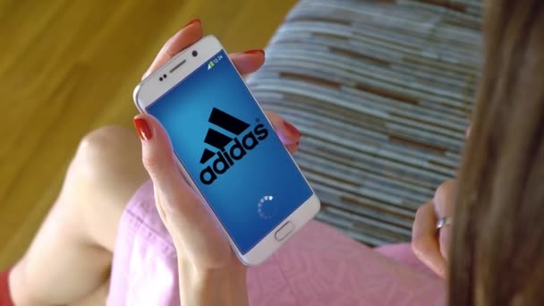 Young woman holding a cell phone with loading Adidas mobile app. Conceptual editorial 4K clip — Stock Video