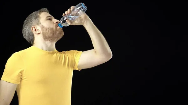 Thirsty gray haired man in yellow tshirt drinking water from small pastic bottle. Black background — Stock Photo, Image