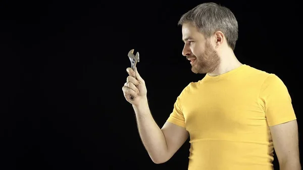 Hombre alegre en camiseta amarilla sostiene llave ajustable. Conceptos de bricolaje, reparación, construcción amateur o mejora del hogar. Fondo negro — Foto de Stock