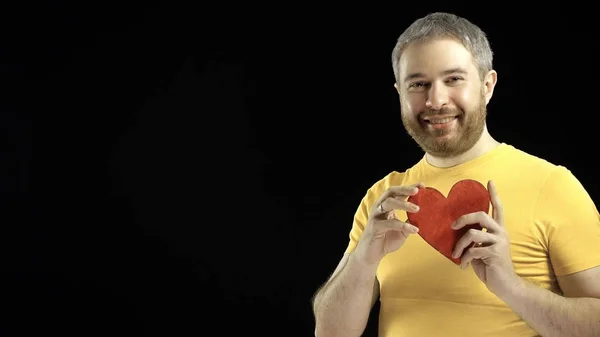 Hombre guapo en camiseta amarilla tiene forma de corazón rojo. Amor, romance, citas, conceptos de relación. Fondo negro — Foto de Stock