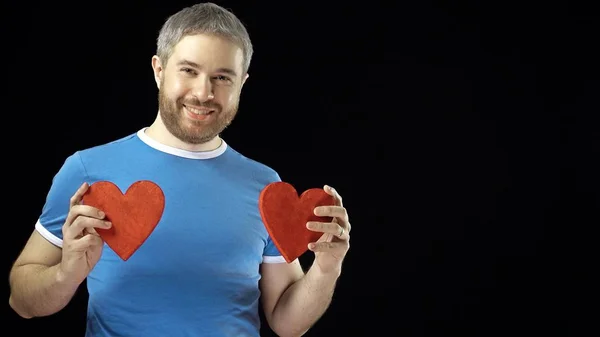 Hombre feliz en camiseta azul tiene dos formas de corazón rojo. Amor, romance, citas, conceptos de relación. Fondo negro — Foto de Stock