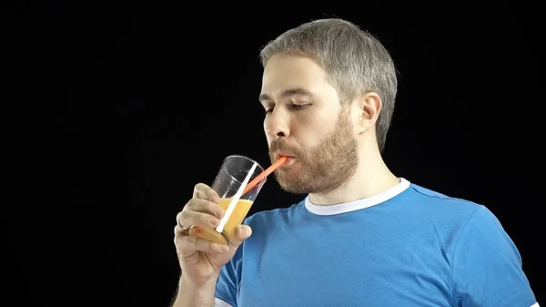 Homem de cabelos grisalhos barbudo atlético em camiseta azul bebendo suco de laranja com uma palha. Fundo preto — Fotografia de Stock