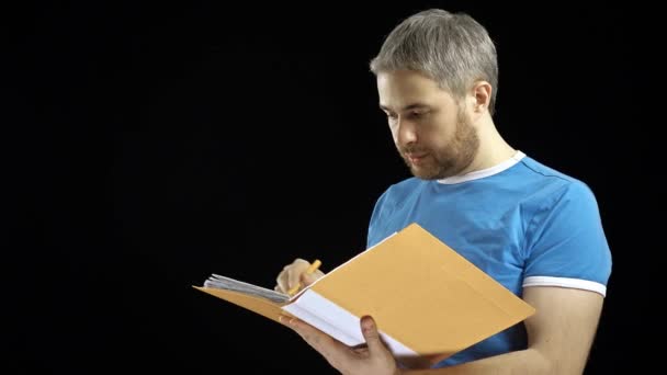 Hombre guapo en camiseta azul mirando a través de papeles en carpeta amarilla. Cotracto, facturas, conceptos de lista de verificación. Fondo negro. Vídeo 4K — Vídeos de Stock