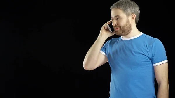 Alegre hombre guapo en camiseta azul habla en su teléfono celular sobre fondo negro — Foto de Stock