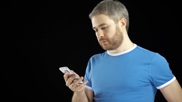 Ein gutaussehender Mann im blauen T-Shirt tippt auf seinem Smartphone-Touchscreen vor schwarzem Hintergrund. 4k-Video — Stockvideo