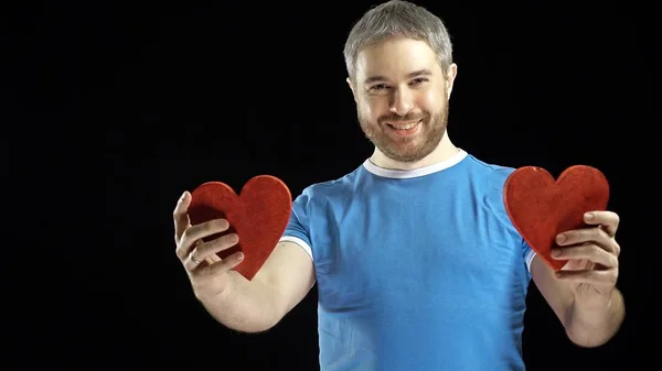 Hombre barbudo sonriente en camiseta azul tiene dos formas de corazón rojo. Amor, romance, citas, conceptos de relación. Fondo negro — Foto de Stock