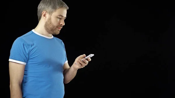 Schöner bärtiger Mann im blauen T-Shirt mit seinem Handy vor schwarzem Hintergrund — Stockfoto