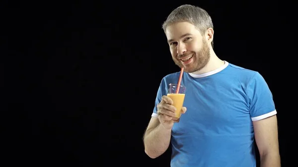 Бородатый молодой человек в синей футболке пьет апельсиновый сок с соломинкой. Чёрный фон — стоковое фото
