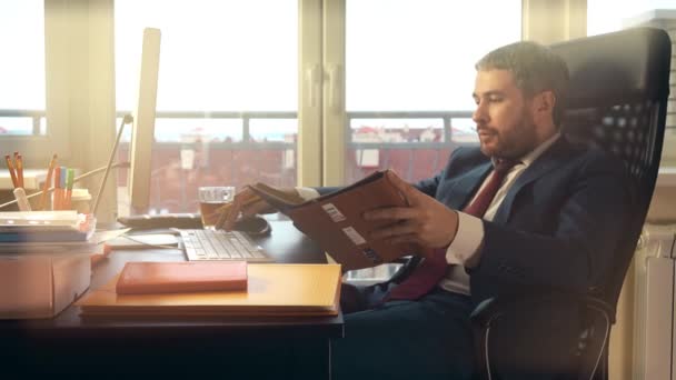 Joven barbudo trabajando en su computadora y escribiendo en su cuaderno en la oficina. Vídeo 4K — Vídeos de Stock