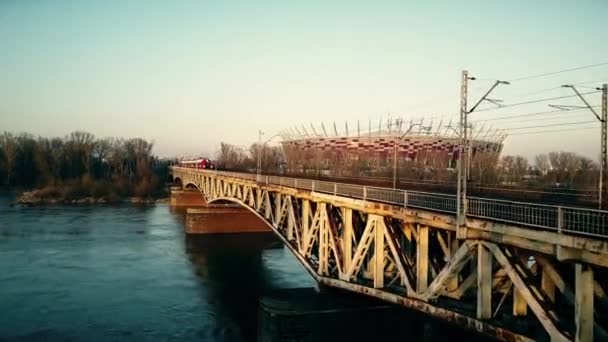 GARANTIA, POLÓNIA - MARÇO, 27, 2017. Tiro aéreo de trem de passageiros vermelho que se move na ponte ferroviária contra a arena moderna. Vídeo 4K — Vídeo de Stock