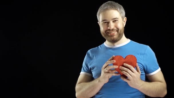 Улыбающийся бородатый мужчина в синей футболке, придающий красную форму сердцу. Любовь, романтика, свидания, концепция предложения. Чёрный фон. 4K видео — стоковое видео