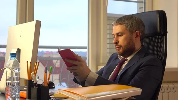 Handsome bearded businessman working on his computer and writing in his red notebook at the office — Stock Photo, Image