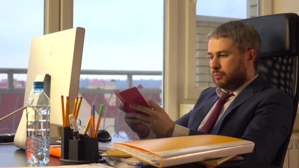 Schöner bärtiger Geschäftsmann arbeitet an seinem Computer und schreibt in sein rotes Notizbuch — Stockvideo