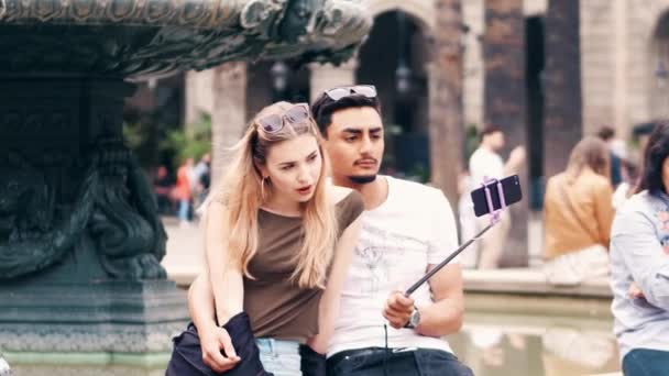 BARCELONA, ESPANHA - 16 de abril de 2017. Belo casal jovem interracial fazendo selfies engraçadas com um telefone celular e uma vara perto da fonte. Vídeo 4K — Vídeo de Stock