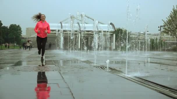 Plan au ralenti d'une jeune femme heureuse de race mixte courant sous la pluie près des fontaines du parc de la ville — Video