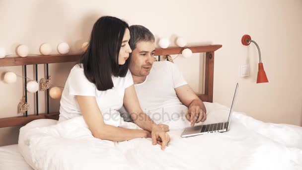 Счастливая пара совершает покупки онлайн, используя ноутбук в постели дома — стоковое видео