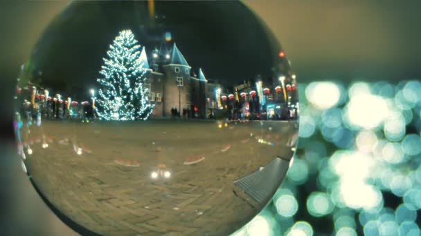 Unknown tourists walk on the square decorated for Christmas and New Year in the evening, view through the glass ball. Amsterdam, Netherlands — Stock Video