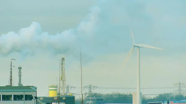 Вращающийся ветрогенератор и дым над промышленной зоной. Концепции производства чистой и грязной энергии — стоковое фото
