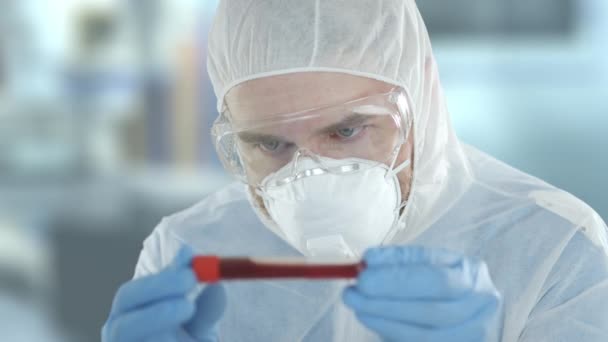 El asistente de laboratorio preocupado que usa traje protector examina la sangre o la muestra de líquido rojo en el tubo de laboratorio. Disparo en la cámara roja — Vídeo de stock