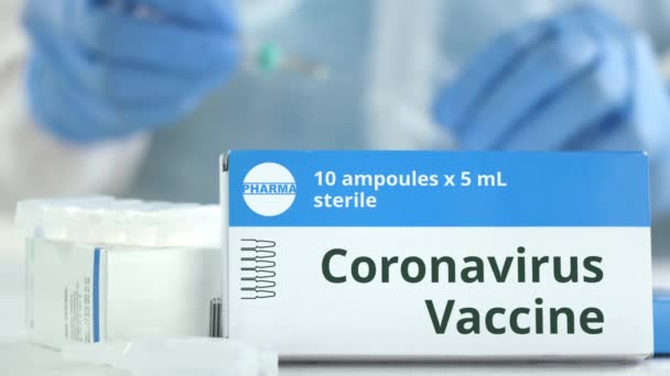 Coronavirus vakcinát tartalmazó doboz az asztalon homályos laborasszisztens ellen. Fiktív gyógyszerészeti logó — Stock videók