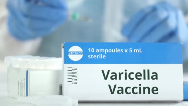 Scatola con vaccino contro la varicella sul tavolo contro l'assistente di laboratorio offuscata o il medico. Logo farmaceutico fittizio — Video Stock