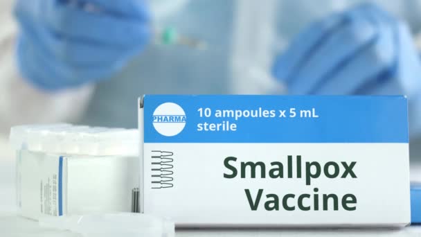 Doos met pokken vaccin op tafel tegen wazig lab assistent. Fictief phaceutisch logo — Stockvideo